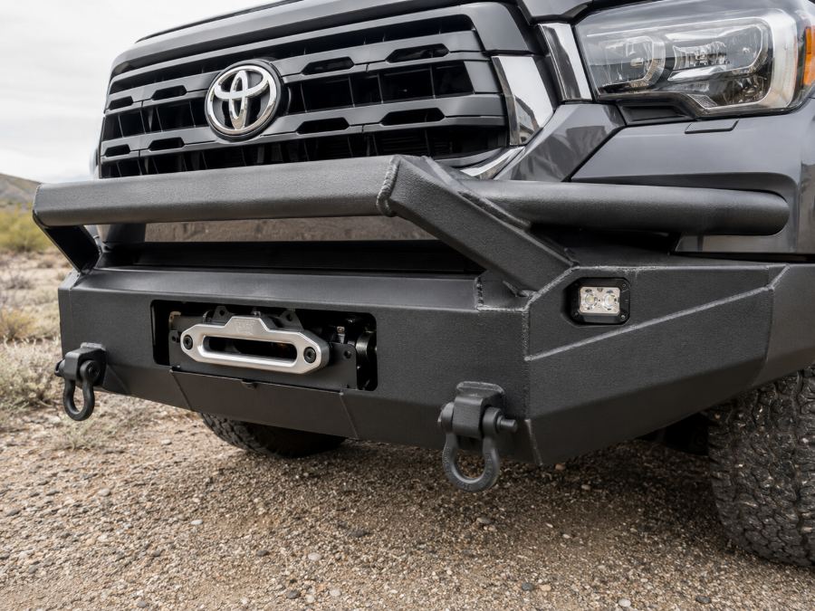 Heavy-duty steel front bumper with winch, fairlead, recovery shackles and integrated fog light in textured black powder coat.