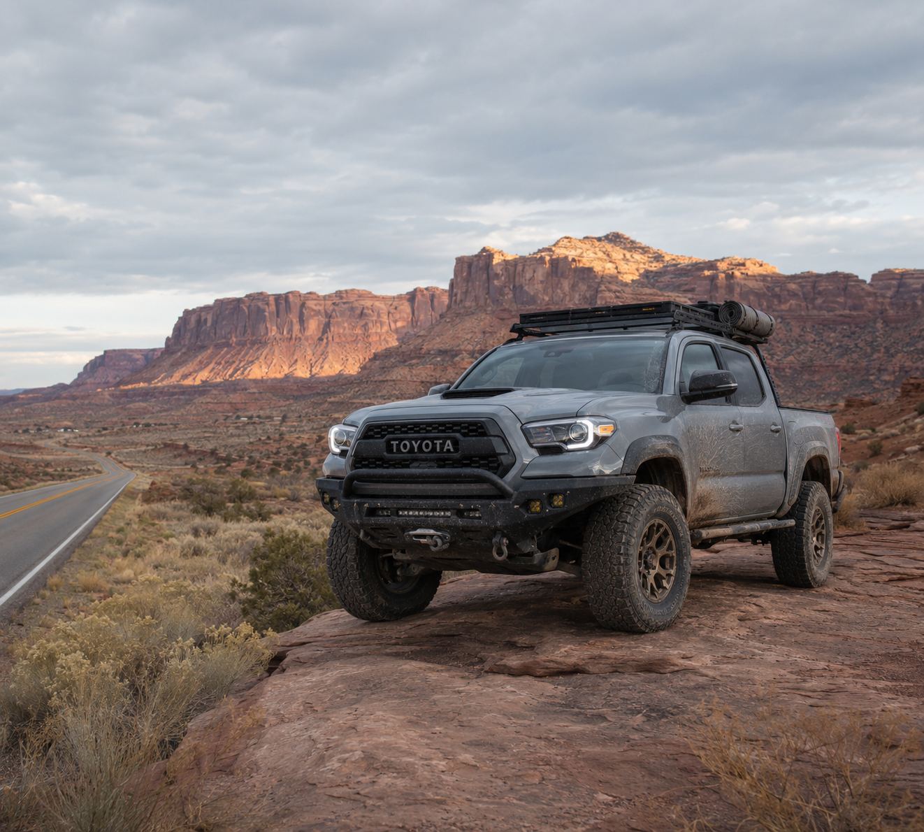 Pickup truck fitted with a Heavy Path Auto steel front bumper, rock sliders and roof rack, parked on a rock ledge above a desert highway in Utah.