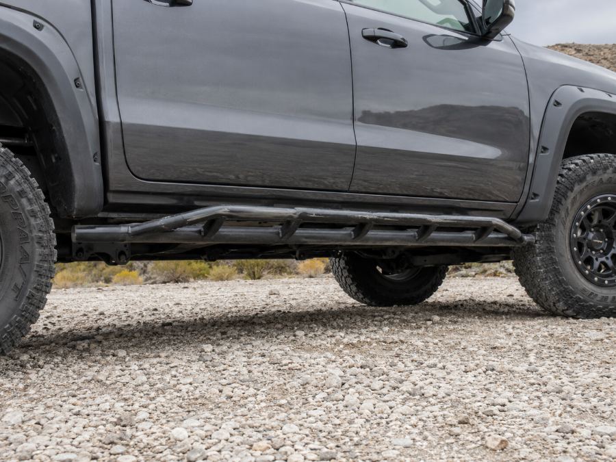 Frame-mounted rock slider in DOM tube running the length of a pickup's rocker panel above gravel.