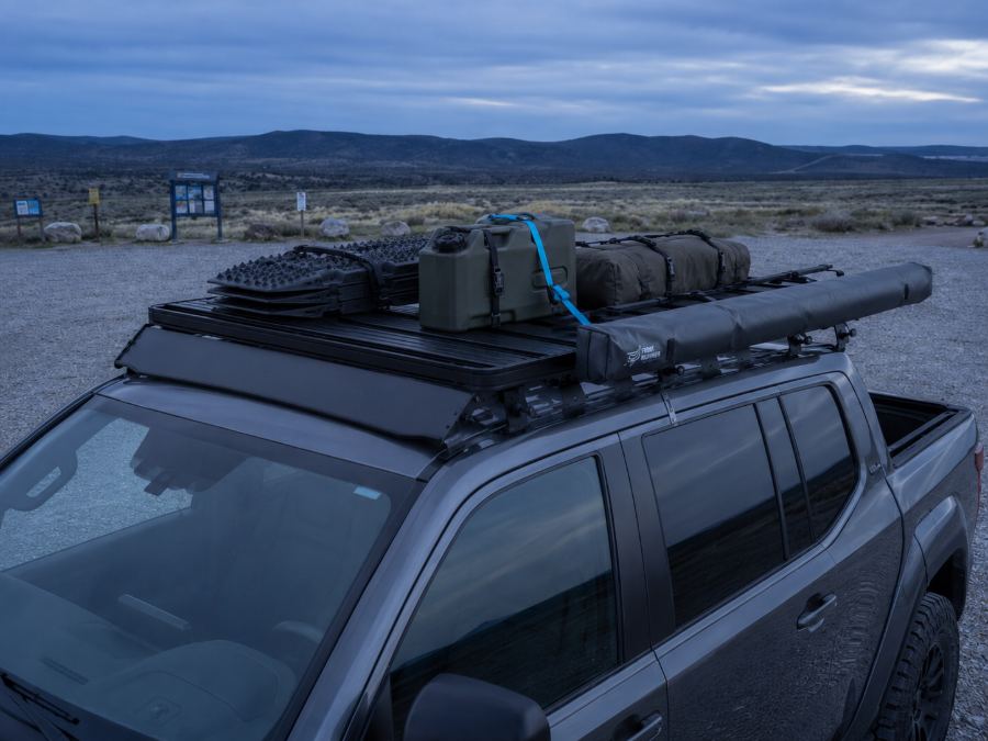 Low-profile roof rack platform on a double-cab pickup loaded with traction boards, a jerry can, a rolled awning and a cyan tie-down strap at blue hour.