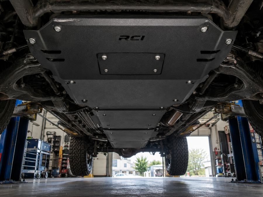 Full underbody skid plate package bolted along a truck frame, seen from below on a shop lift, with a serviceable access panel.